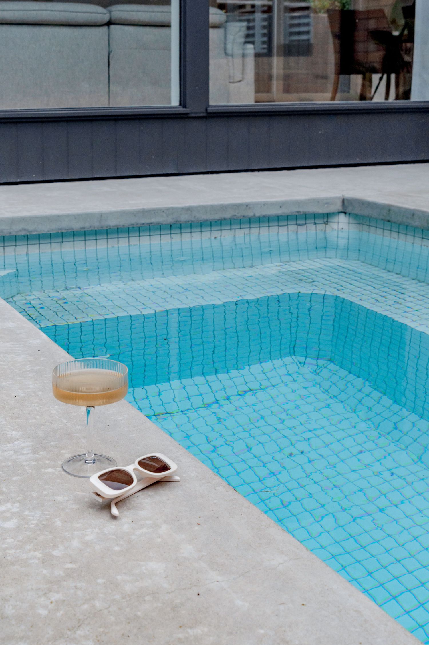 A cocktail glass and white sunglasses rest beside a blue-tiled swimming pool on a concrete ledge.