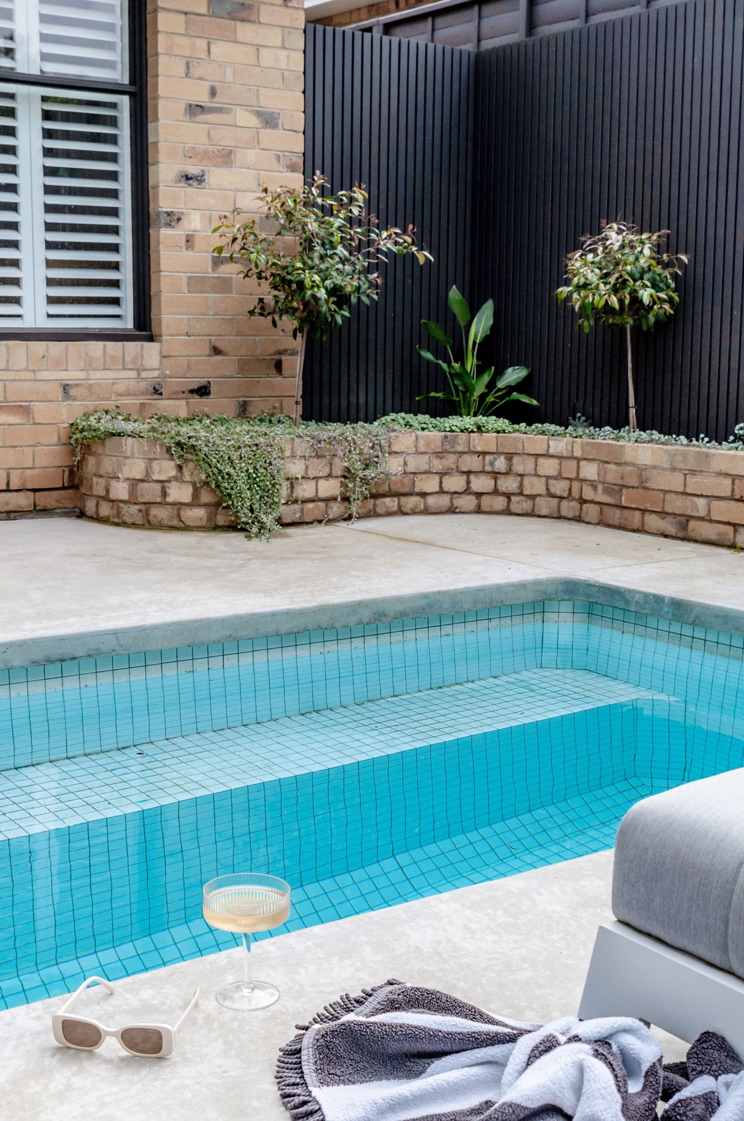 Luxurious backyard pool area with brick retaining wall, potted plants, and lounge accessories beside turquoise water.
