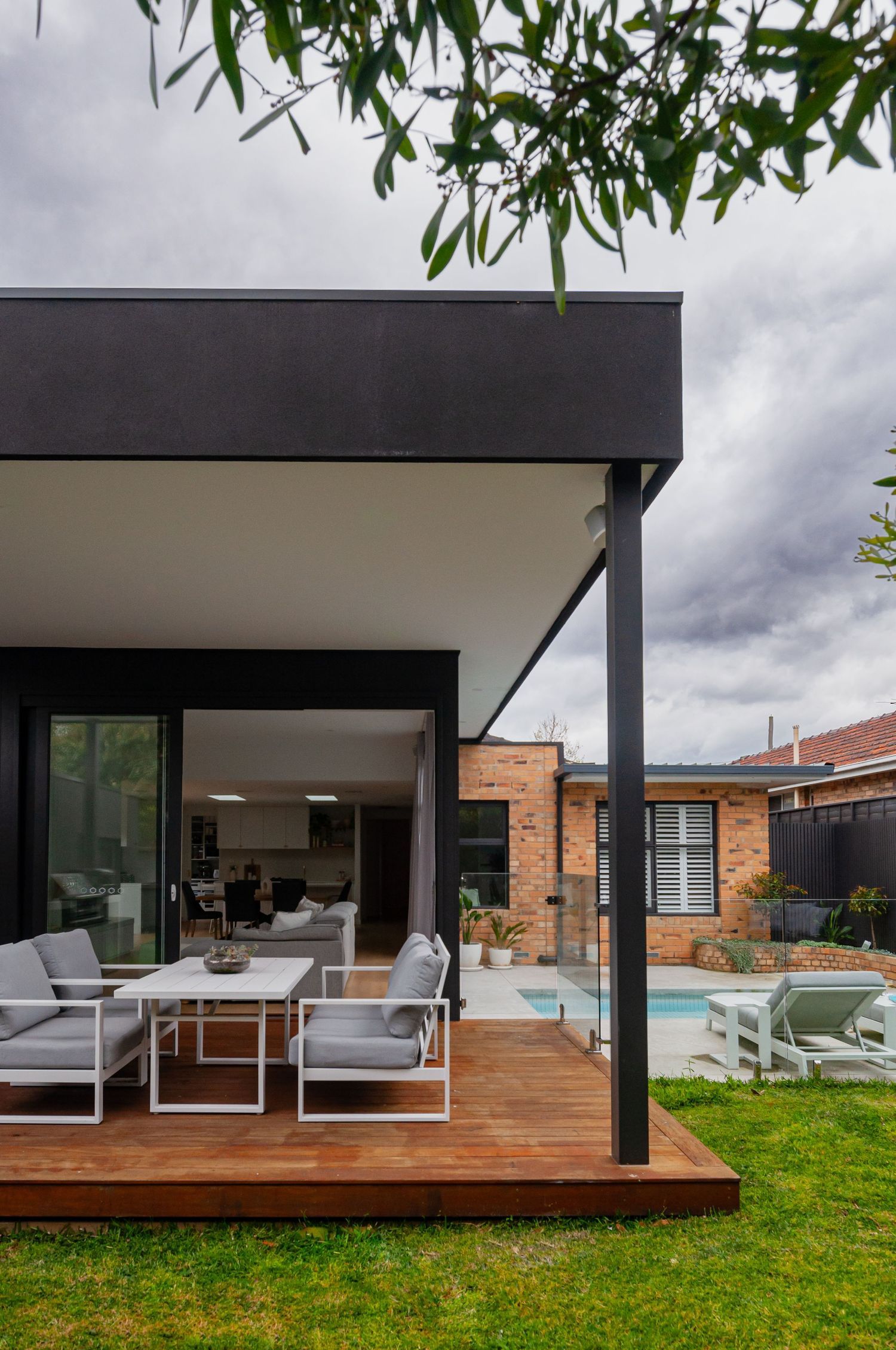 Modern outdoor patio with gray seating and wooden deck overlooking a backyard pool under cloudy skies.