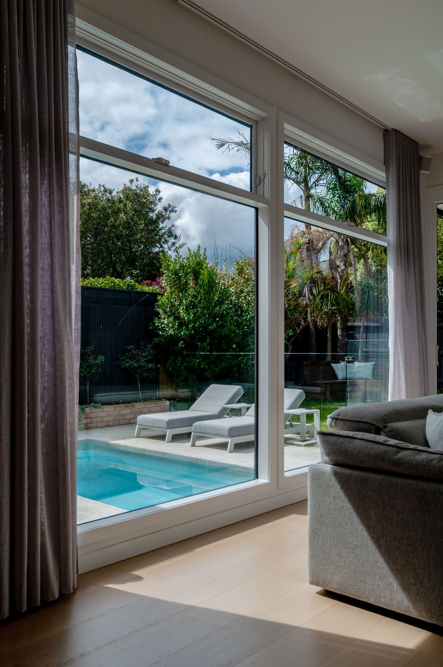 Modern living room with large windows overlooking a pool patio with loungers and tropical landscaping in the backyard.