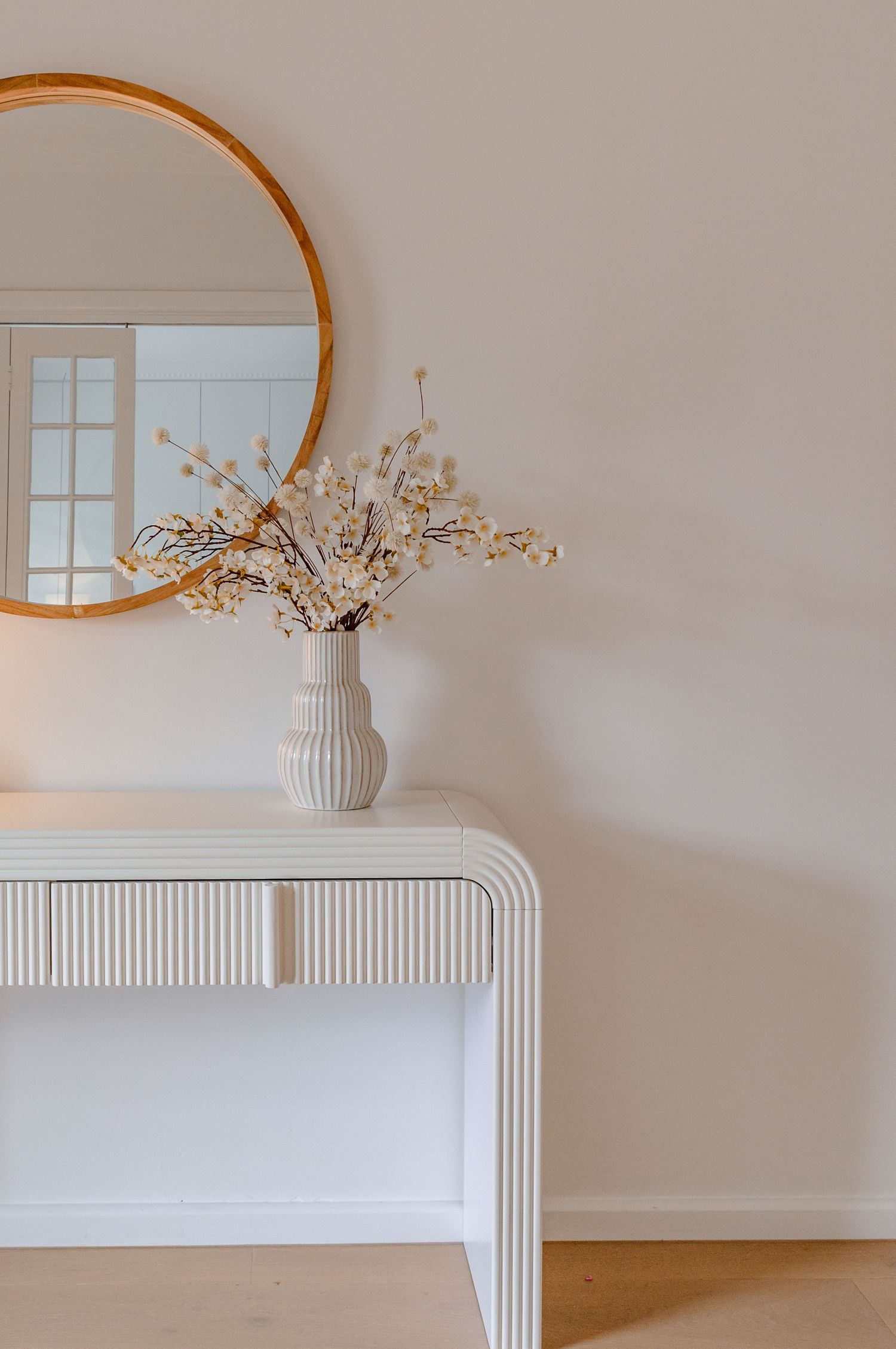 A minimalist white console table with ribbed details paired with a round gold mirror and dried floral arrangement.