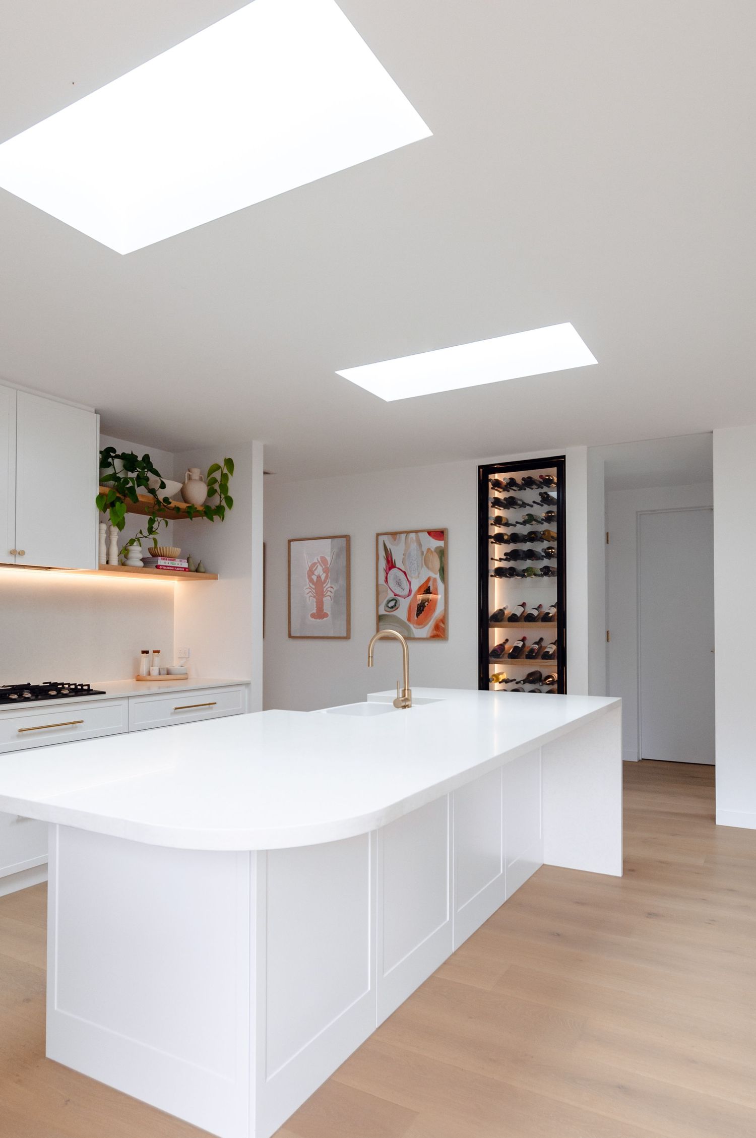 Modern white kitchen with skylight features sleek island, built-in wine storage, and minimalist decor with warm wood floors.