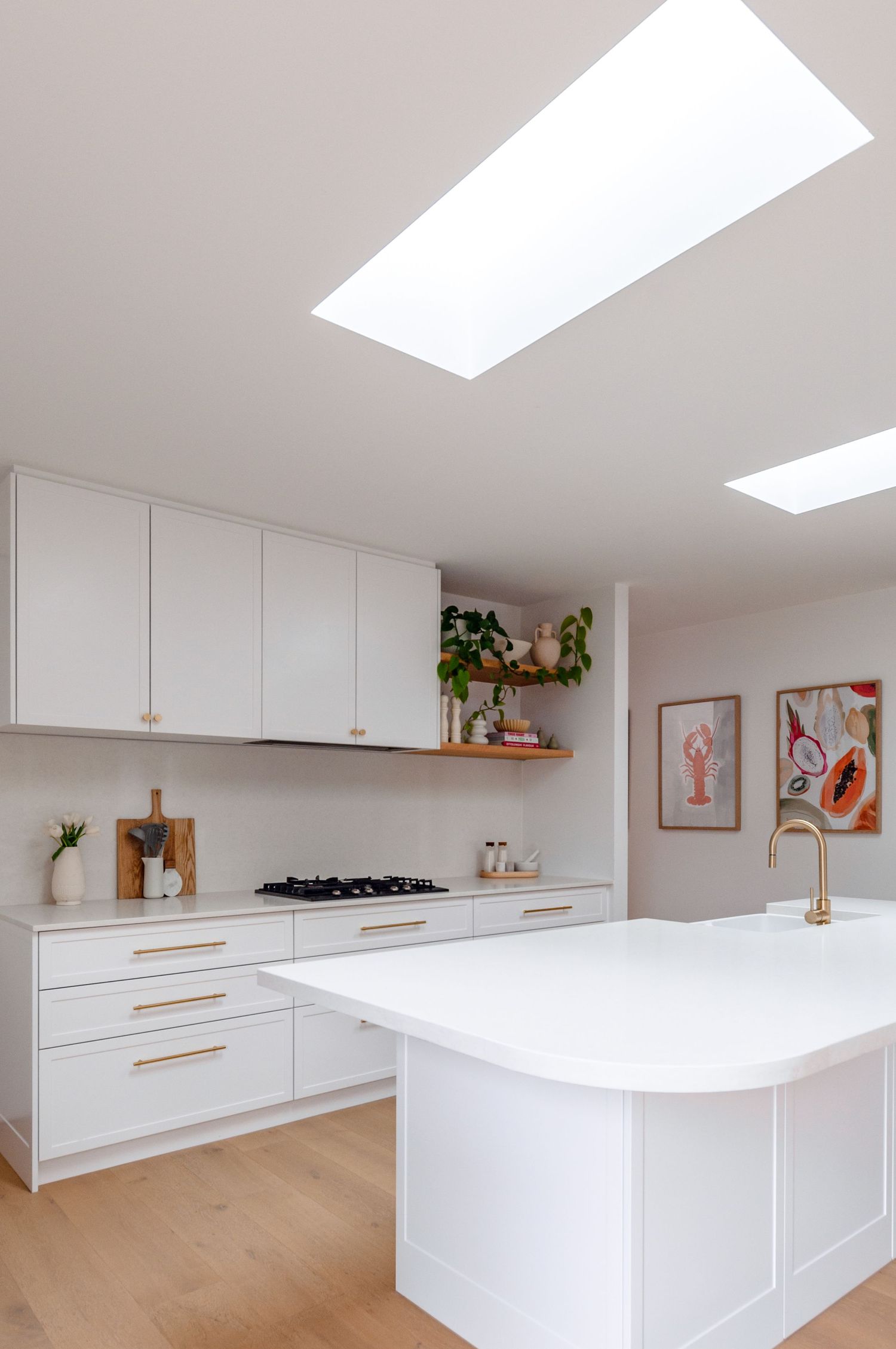 Modern white kitchen with skylight features sleek cabinets, gold hardware, floating shelf, and large center island.