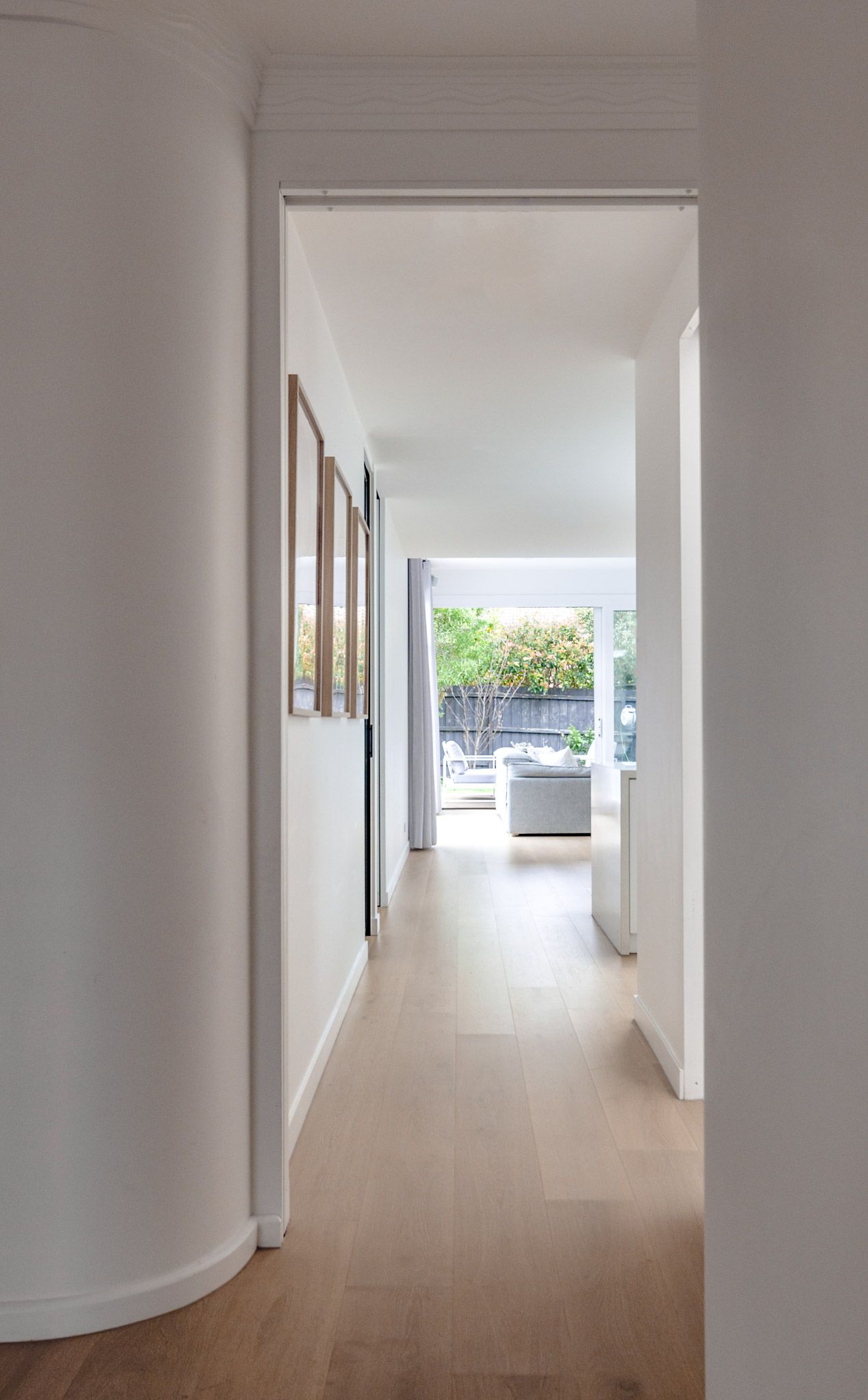 Long bright hallway with light hardwood floors leading to a sun-lit living area with curved wall details.