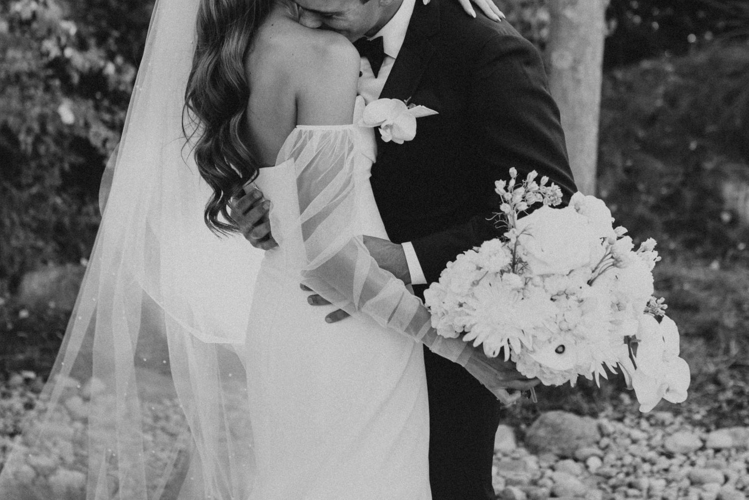 Black and white wedding photograph showing elegant white bridal bouquet and flowing dress details during intimate moment.