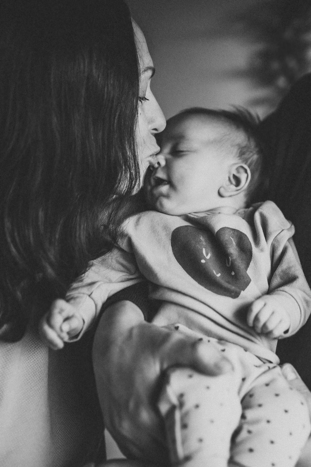 Black and white intimate portrait of mother kissing baby in polka dot outfit.