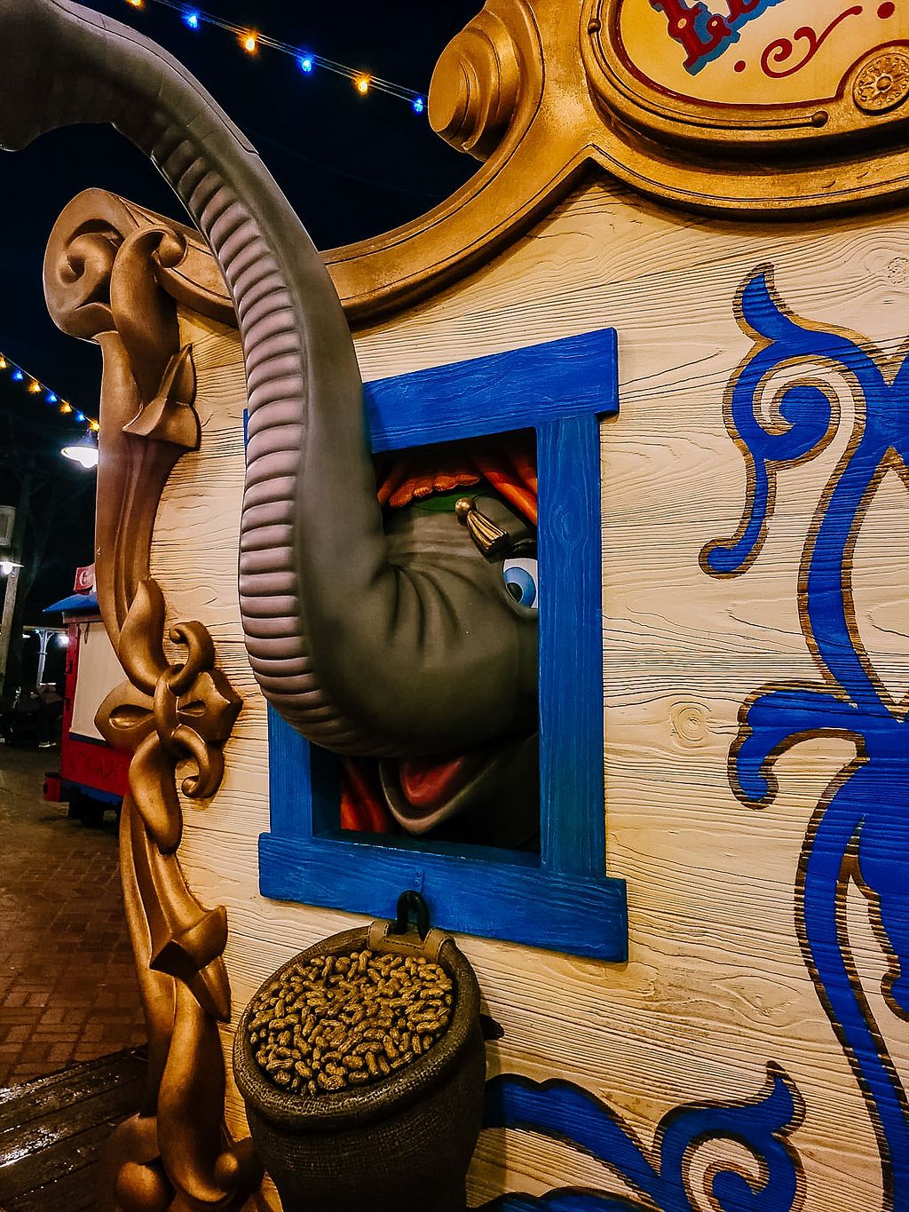 Ornate wooden carnival cart with decorative blue window frame and whimsical gold scrollwork illuminated by string lights at night.