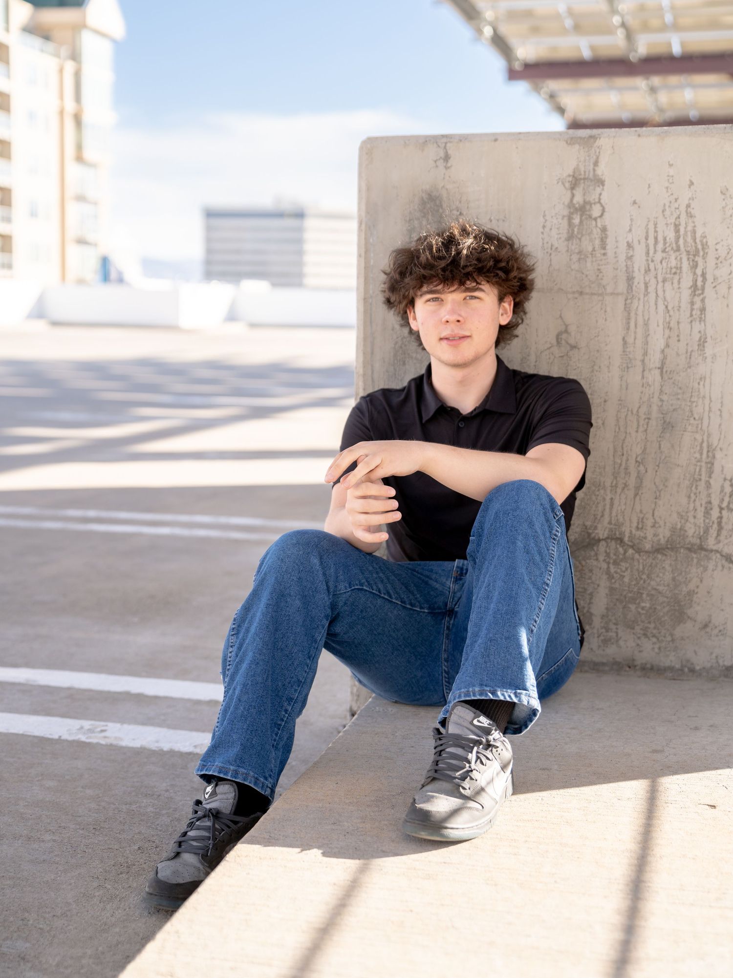 High school senior in a black t-shirt and jeans sitting casually against a concrete wall in an urban setting with sunlight and shadows.