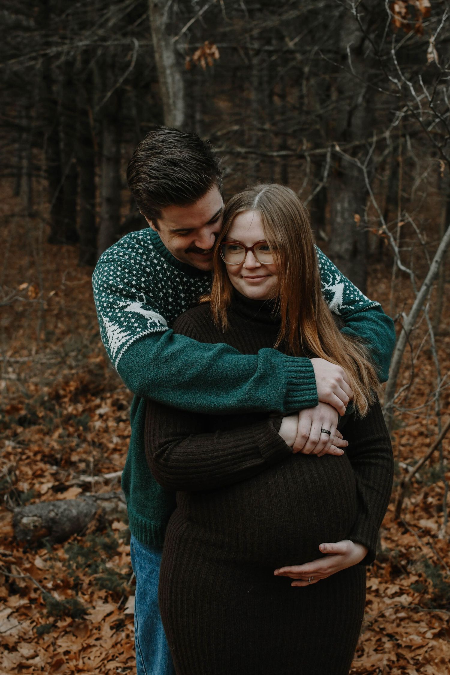 Couple embraces in autumn forest wearing cozy sweaters among fallen leaves and bare trees.