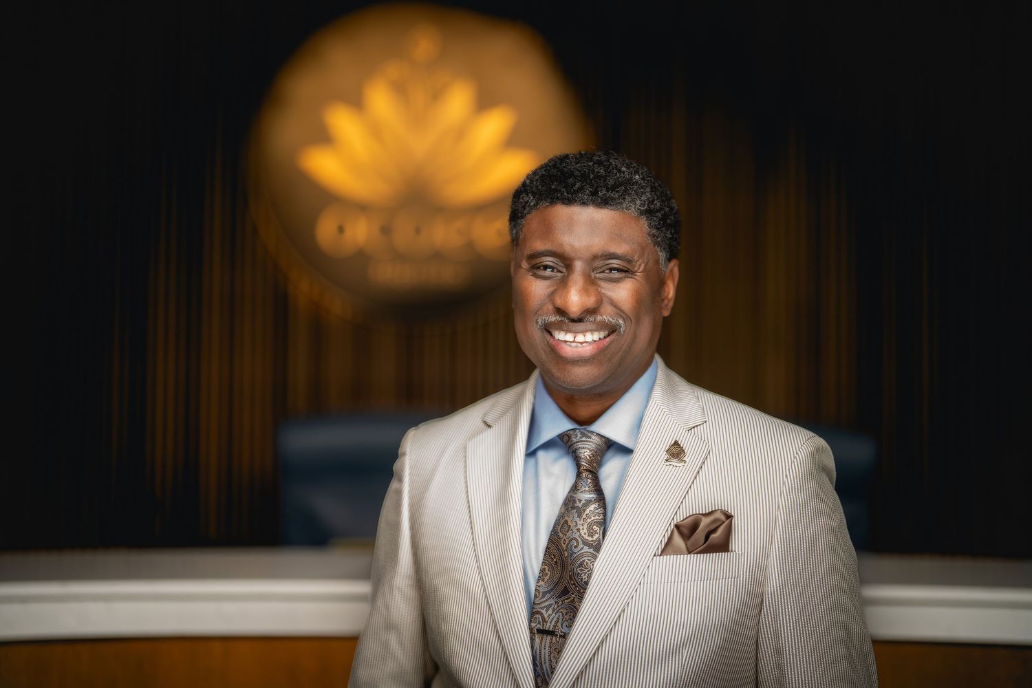 Professional portrait of someone in a light gray suit and patterned tie standing in front of an illuminated logo display.
