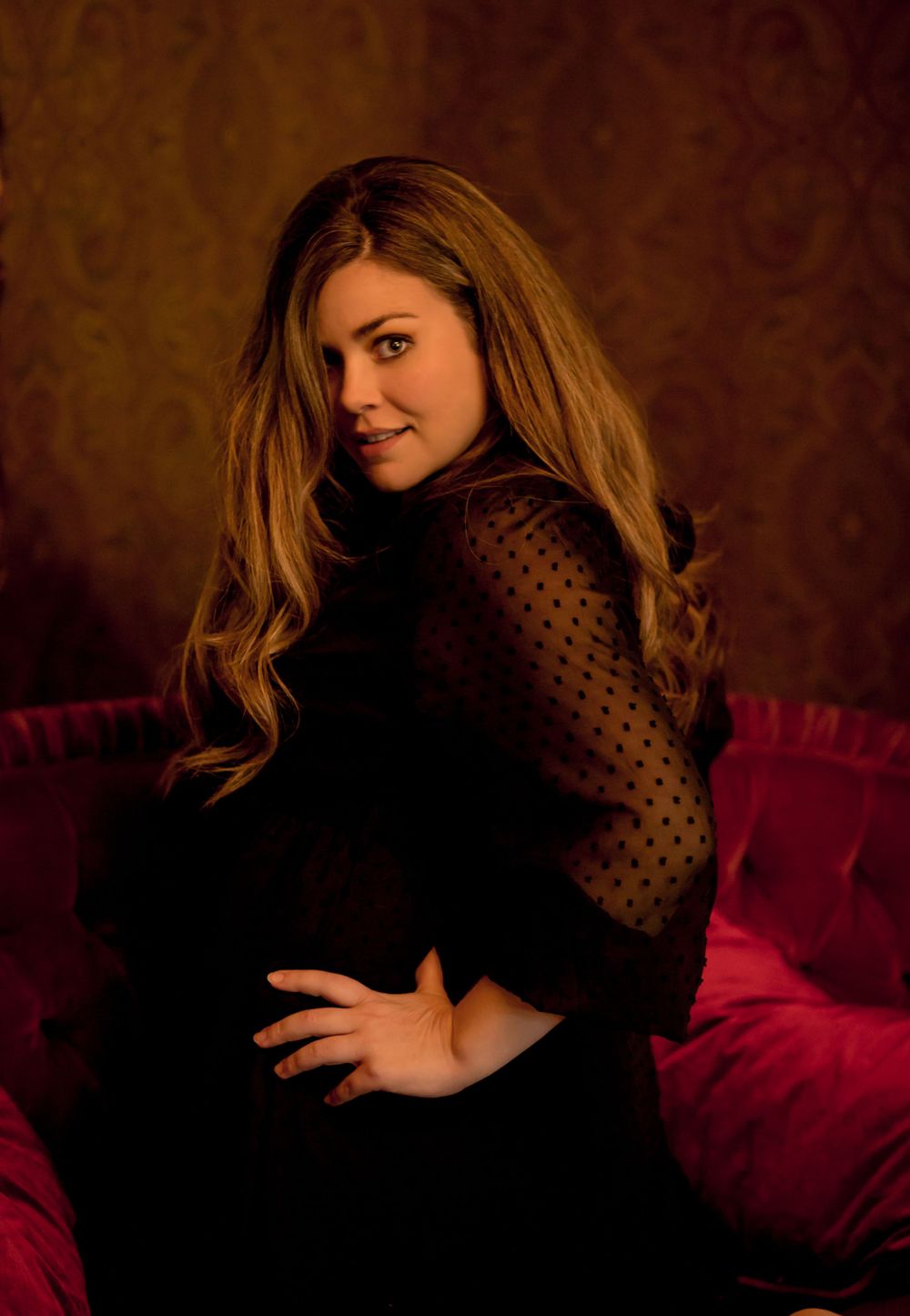Woman with long auburn hair in black polka-dot top seated on red velvet couch against ornate wallpaper background.