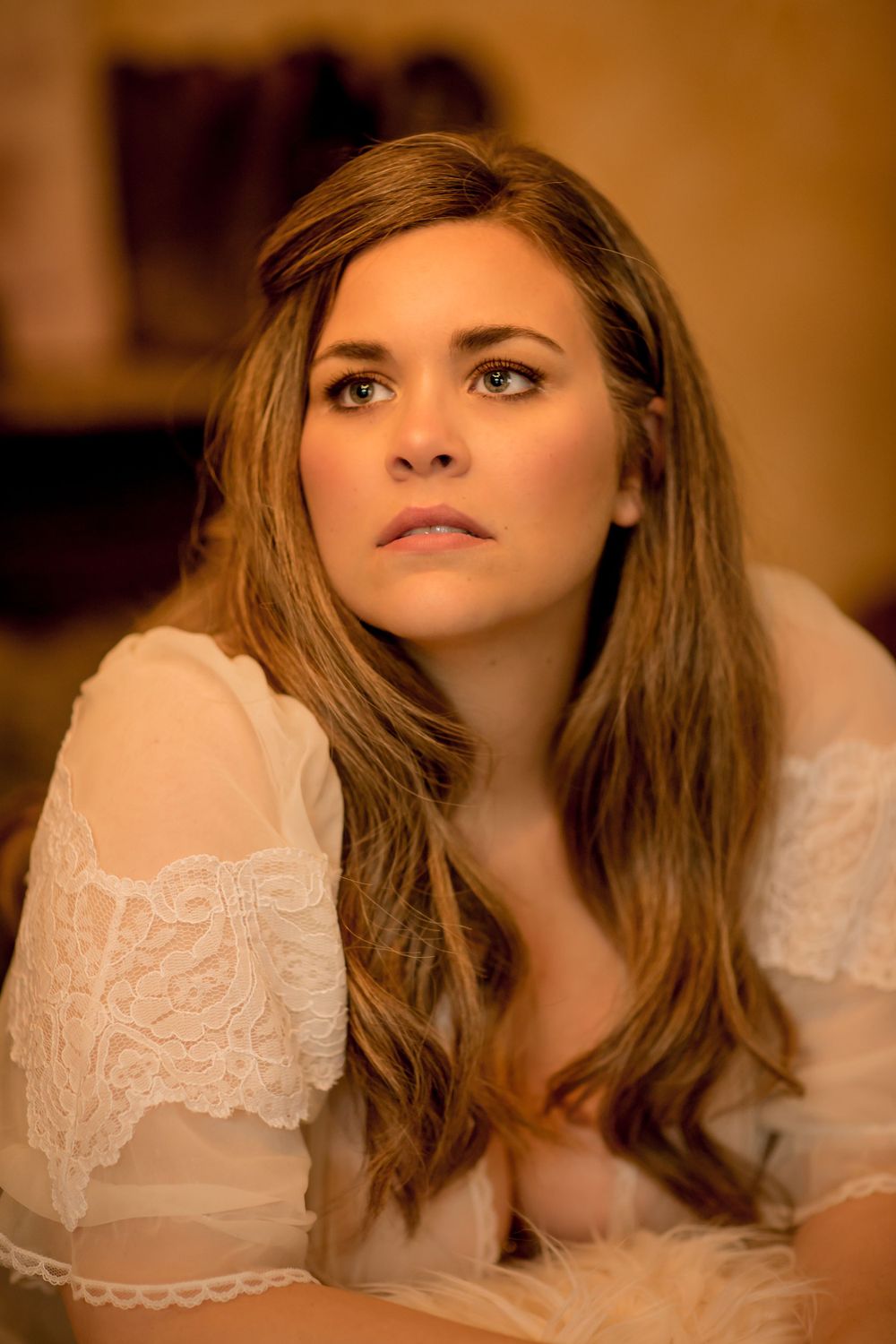 Young woman with long brown hair in white lace top gazing upward with a contemplative expression in warm light.