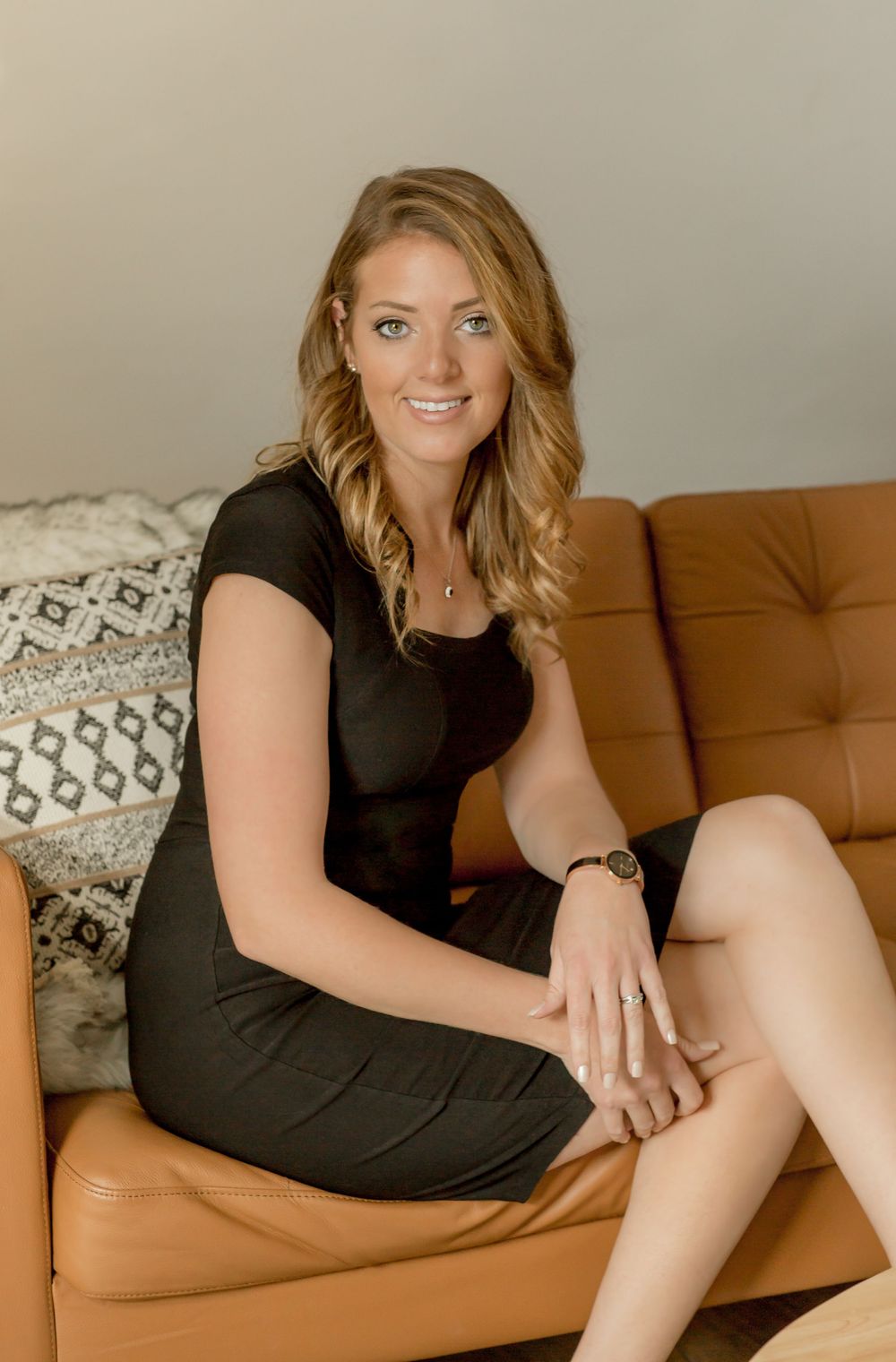 Smiling blonde woman in black dress sitting on tan leather sofa indoors.
