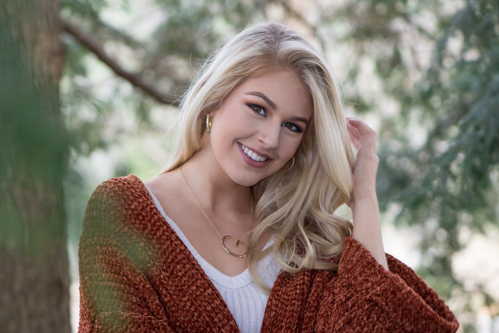 Smiling blonde woman in rust-colored knit cardigan poses outdoors among trees in natural light.