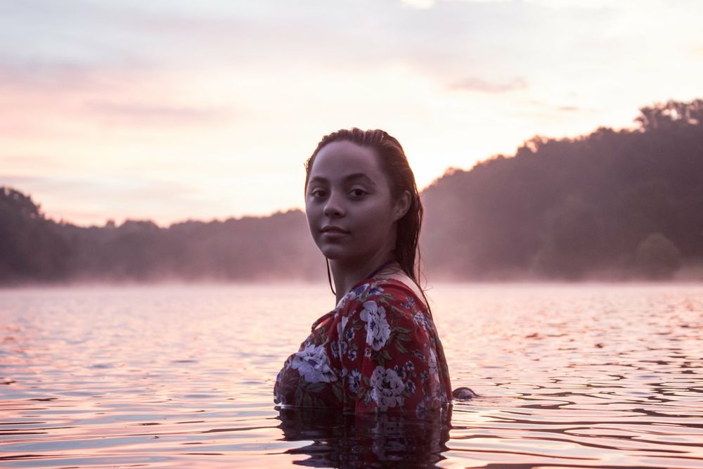Woman in floral top partially submerged in misty lake during pink sunset twilight.