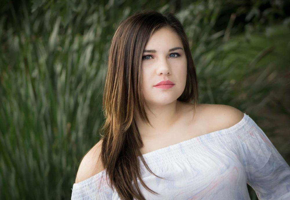 Young woman with long brown hair in white off-shoulder top posing outdoors near greenery.