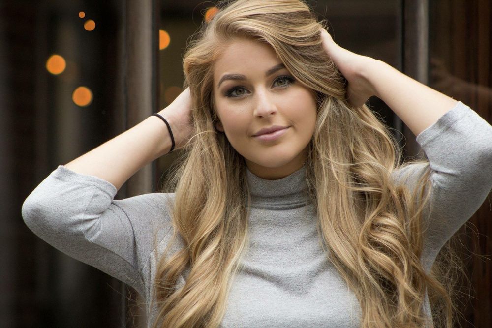 Smiling young blonde woman in gray turtleneck sweater with hands in hair in urban setting.