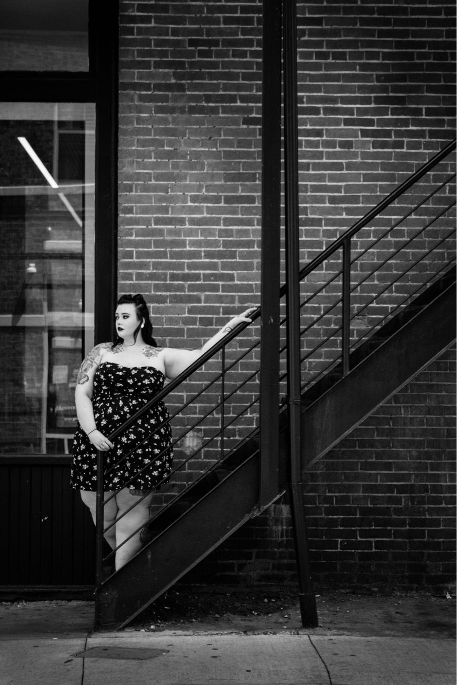 Black and white portrait of tattooed woman in strapless dress leaning on urban fire escape stairs.