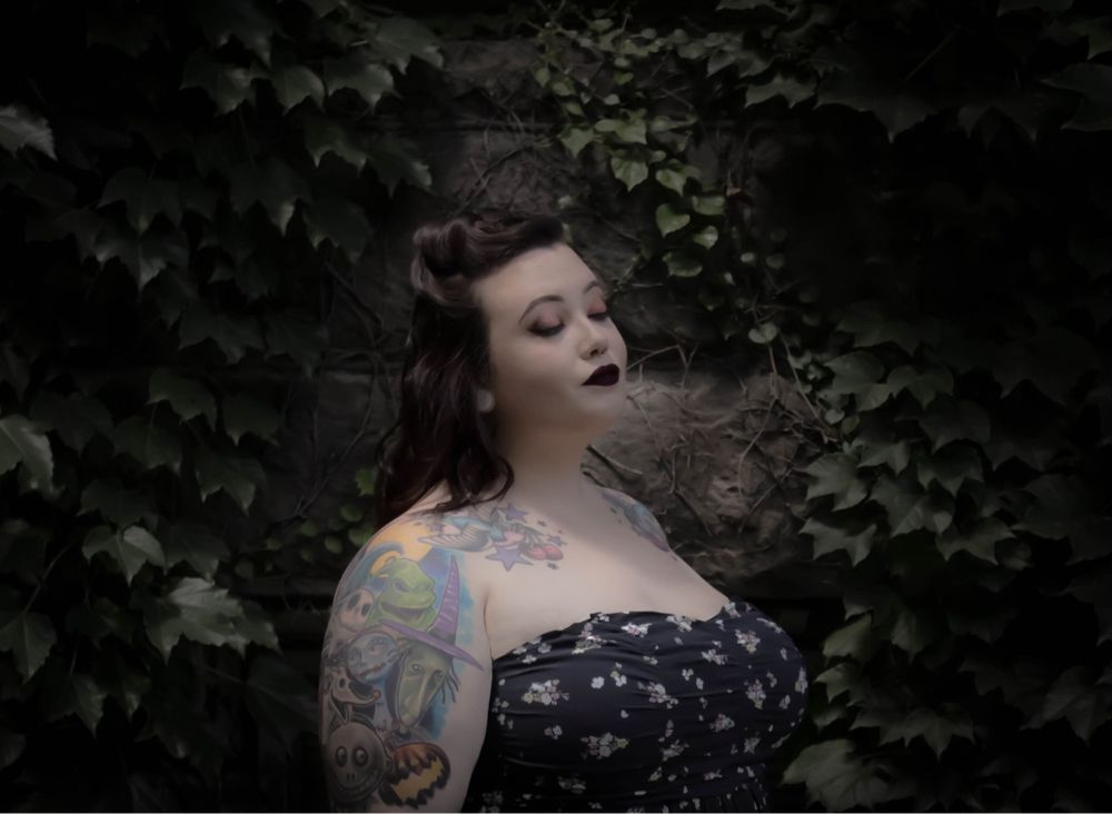 Tattooed woman in floral dress posing dramatically against ivy-covered stone wall in moody lighting.