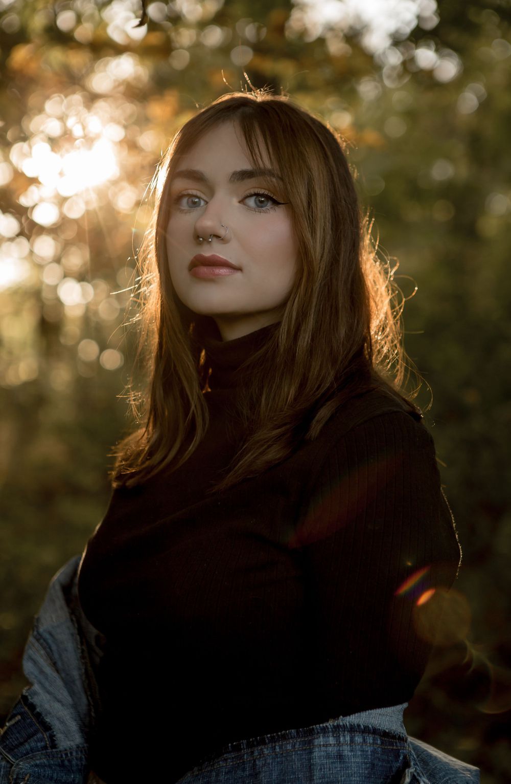 Young woman with auburn hair and nose ring in black turtleneck posed outdoors with golden backlit sunlight through trees.