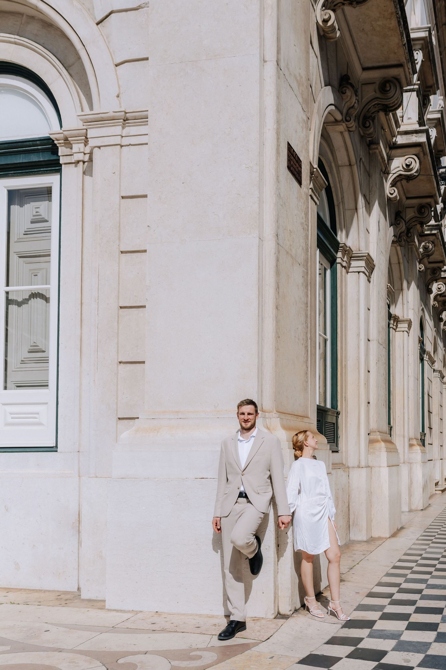 Elegant couple in light-colored outfits walking along ornate architectural building with checkered floor and arched windows.