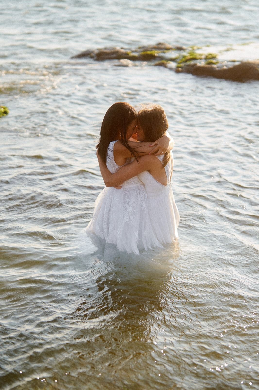 Two people in white wedding dresses embrace while standing in ocean waters during golden hour.