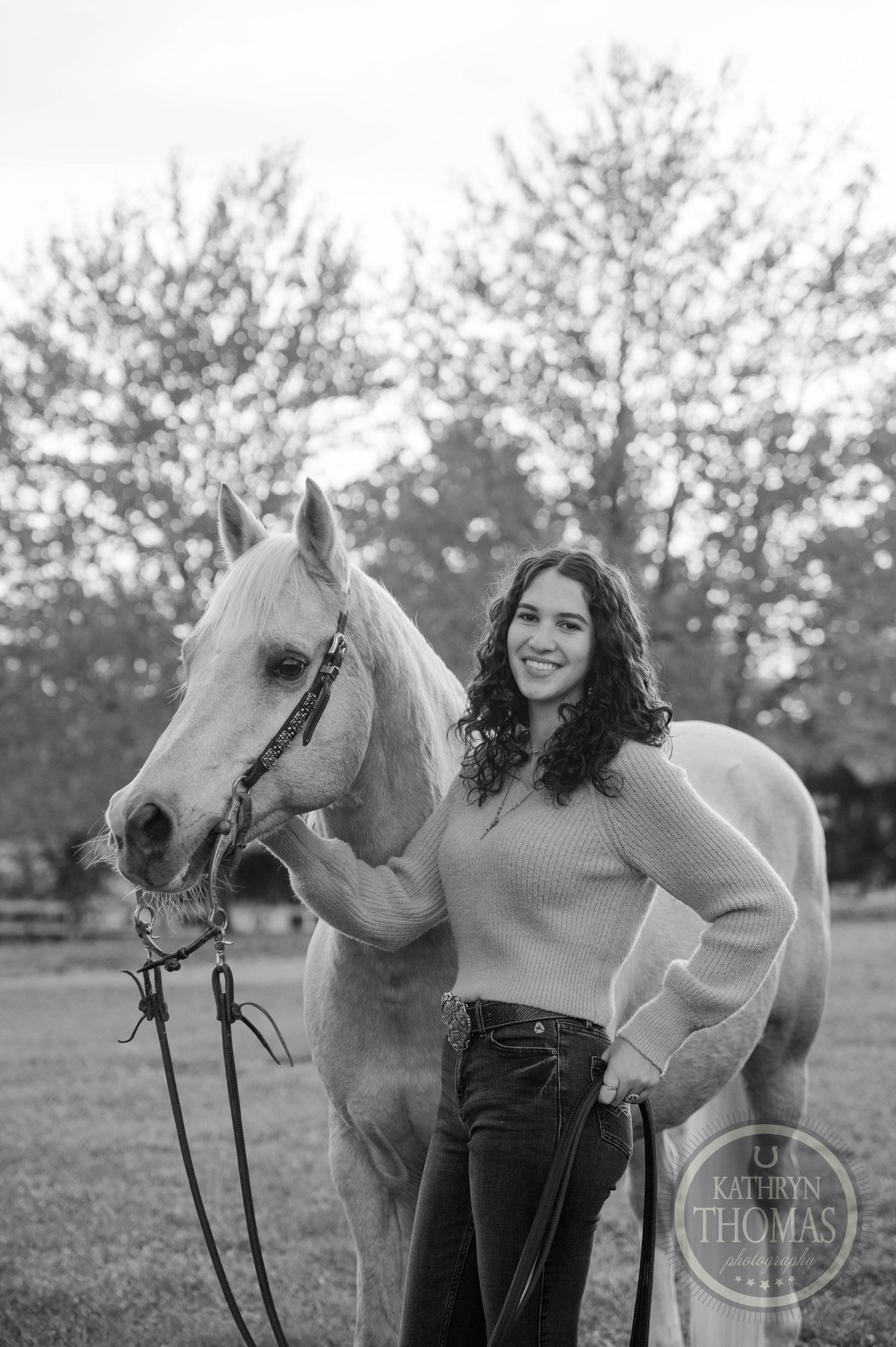 A black and white portrait with someone posing beside a light-colored horse outdoors.