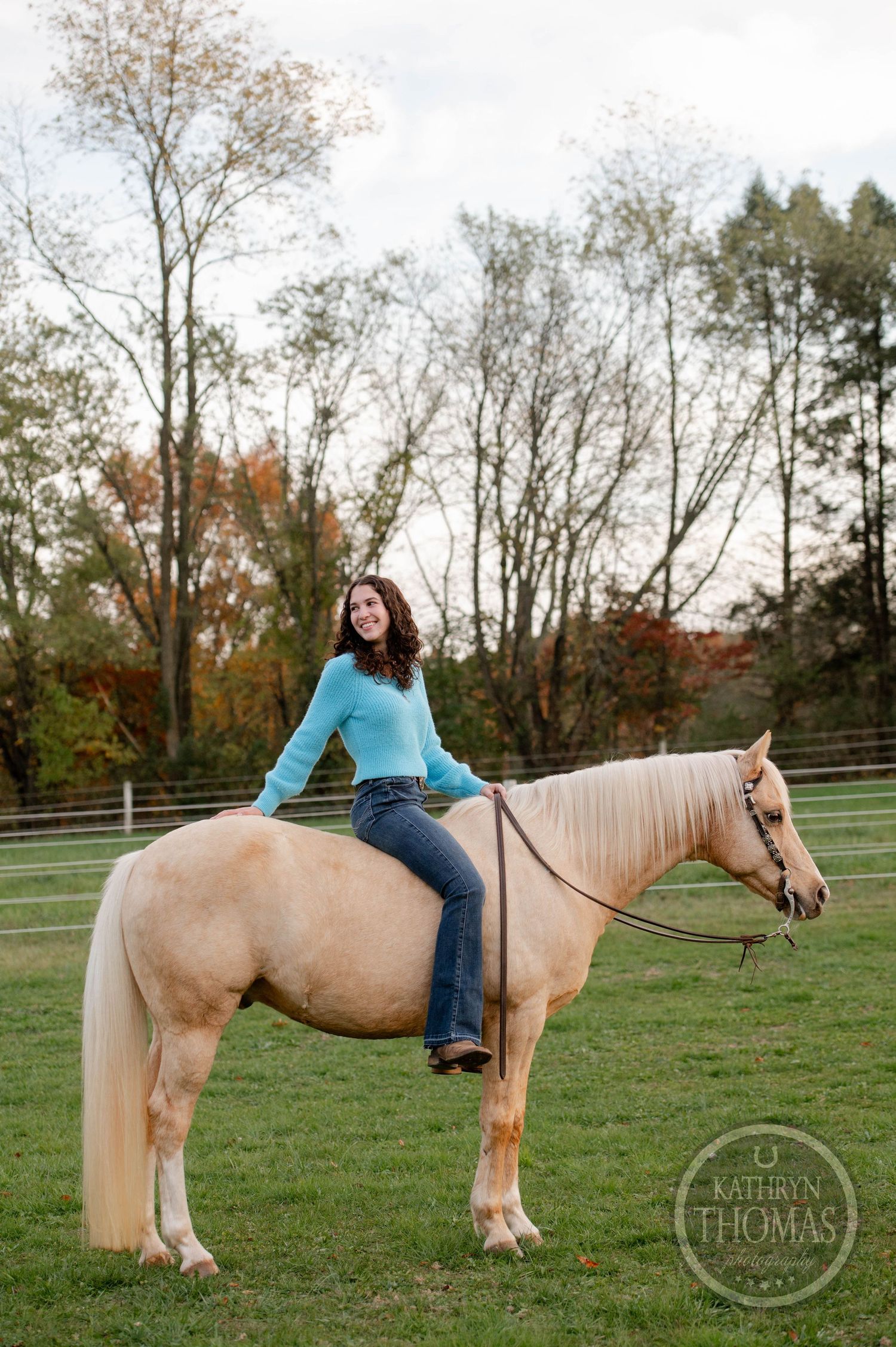 Rider in turquoise sweater sits astride palomino horse in outdoor arena with fall foliage.