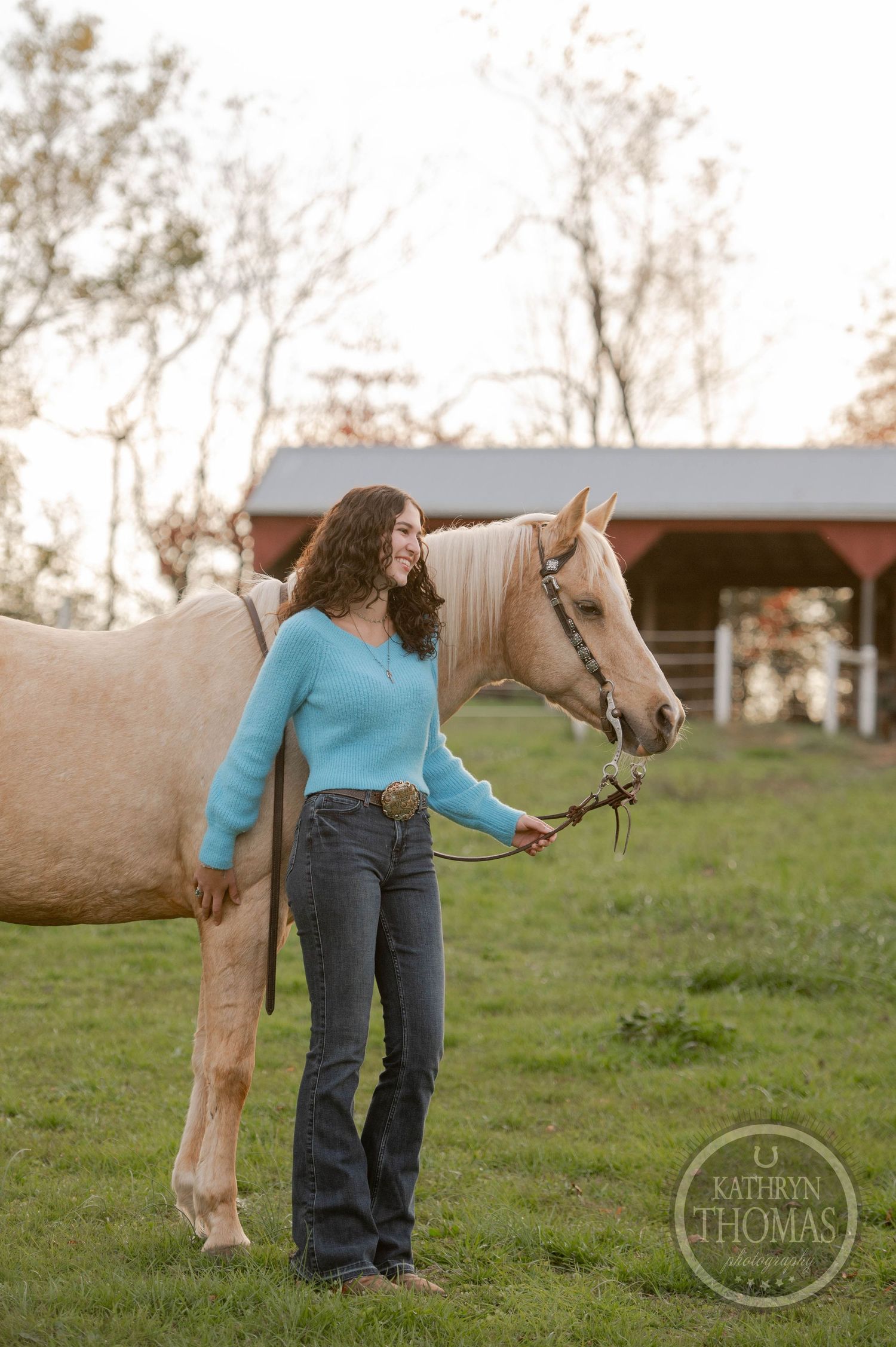 Person in blue sweater and jeans stands with light-colored horse in pasture at sunset.