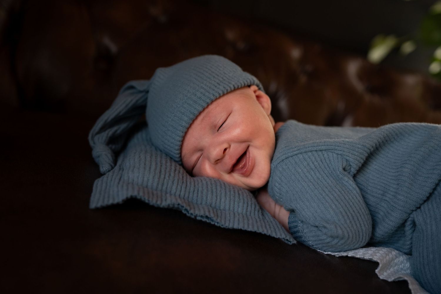 A newborn baby wrapped in soft blue knitwear smiles while sleeping peacefully.