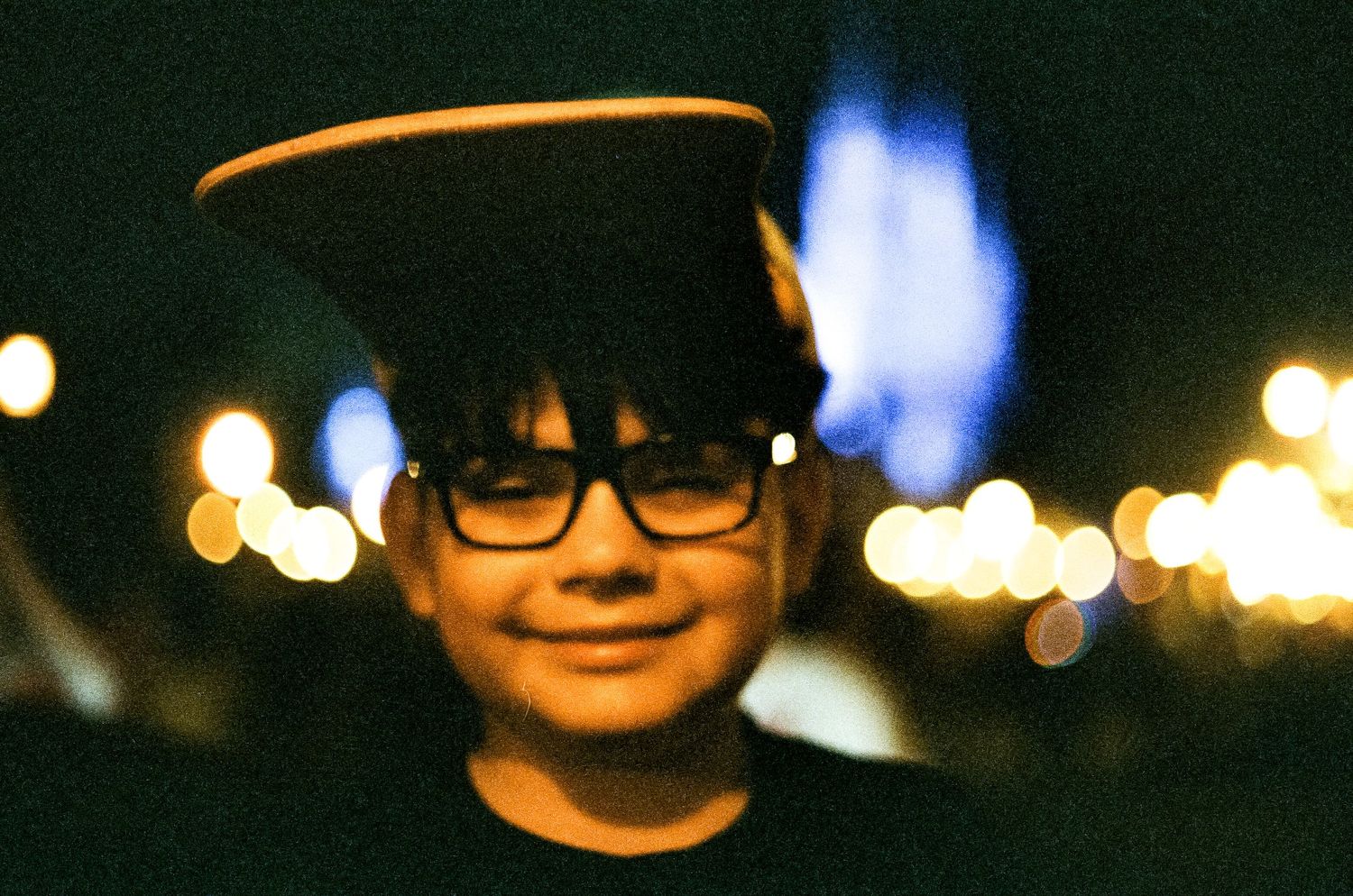 A child wearing glasses smiles at night with blurred city lights glowing in the dark background.