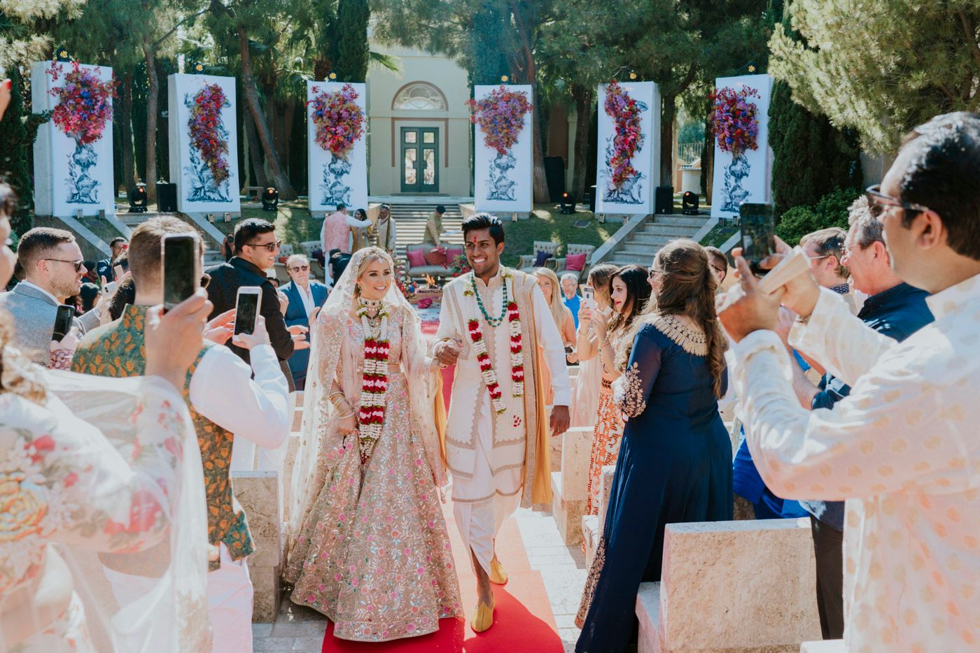 Wedding ceremony guests capture an outdoor celebration with cameras under hanging floral banners on a sunny day.