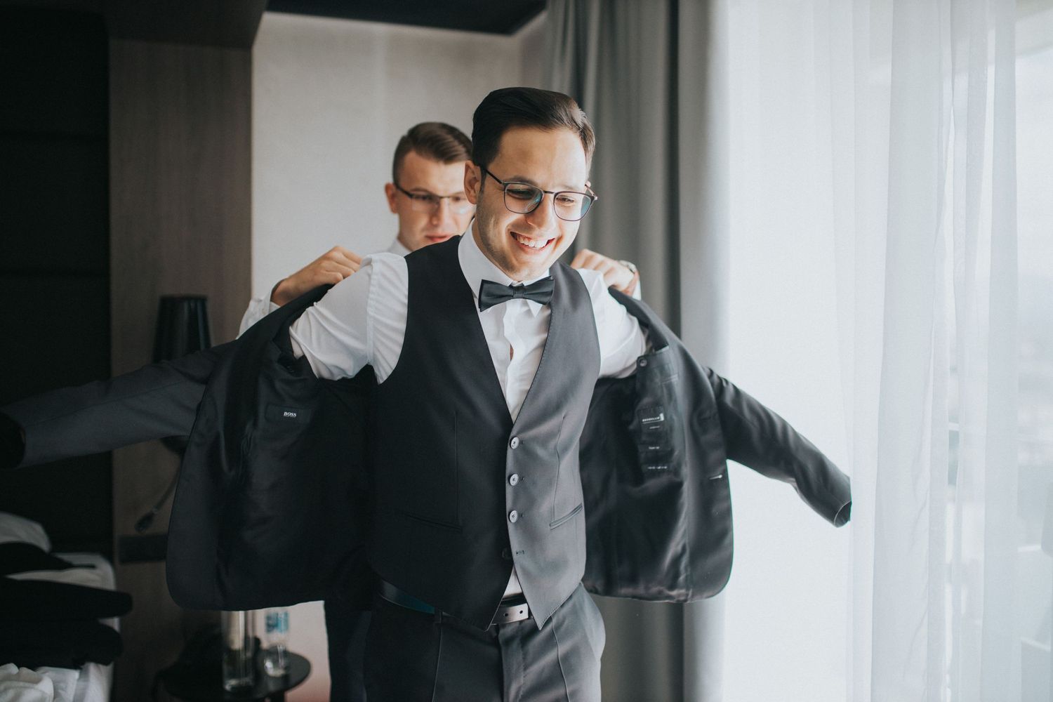 A well-dressed person in a formal vest and bowtie is being helped into a black suit jacket by someone behind them.