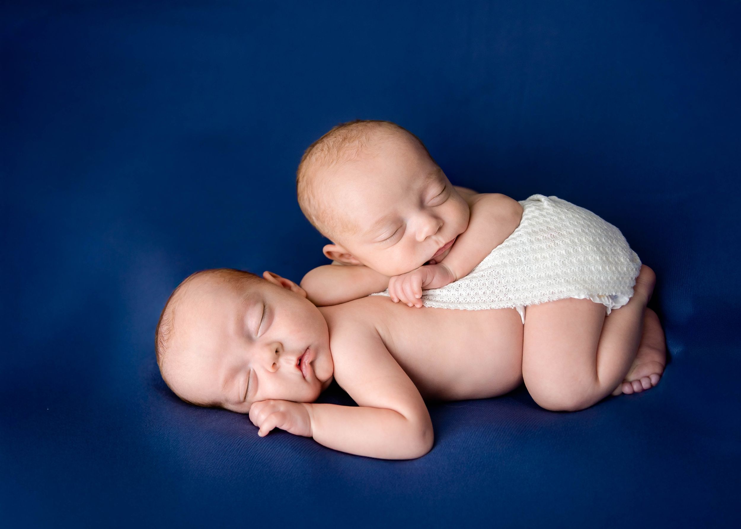 Two sleeping babies in white diapers lying together against a dark blue background. #RoswellNM #TwinNewbornPhotographer #ArtesiaNM #carlsbad