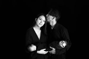 A black and white studio portrait of a couple in dark clothing standing close together against a dark background#TwinNewbornSession #NM