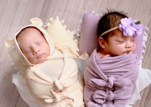 Twin newborns swaddled in cream and lavender wraps lying side by side in a woven basket with floral headband accent. #TwinNewbornPhotography
