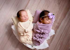 Twin newborns swaddled in cream and lavender wraps lying side by side in a woven basket with floral headband accent. #TwinNewbornPhotography