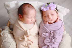 Twin newborns swaddled in cream and lavender wraps lying side by side in a woven basket with floral headband accent. #TwinNewbornPhotography
