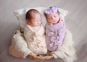 Twin newborns swaddled in cream and lavender wraps lying side by side in a woven basket with floral headband accent. #TwinNewbornPhotography