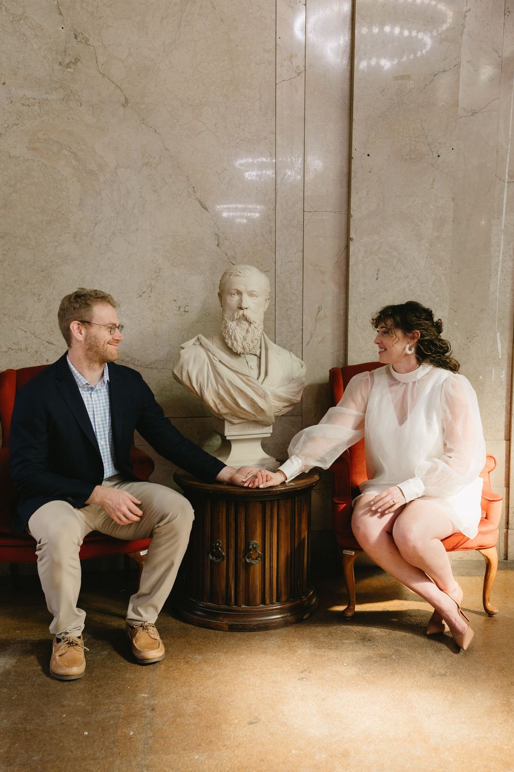 Two people sit conversing near a classical bust sculpture in an elegant interior setting.