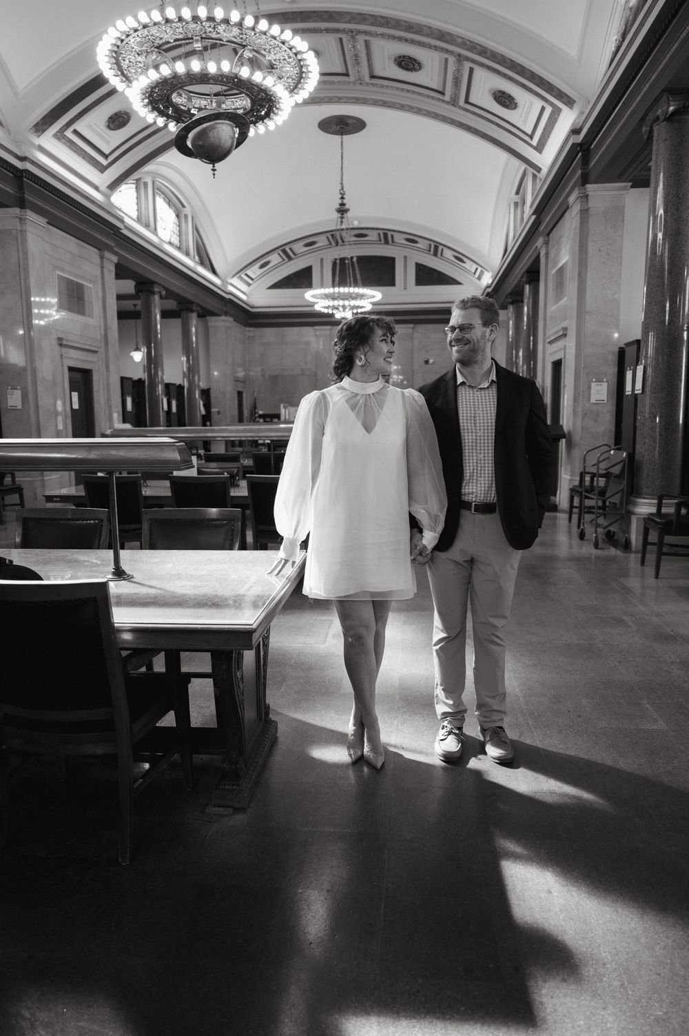 A couple walks through an elegant hallway with ornate chandeliers and architectural details.