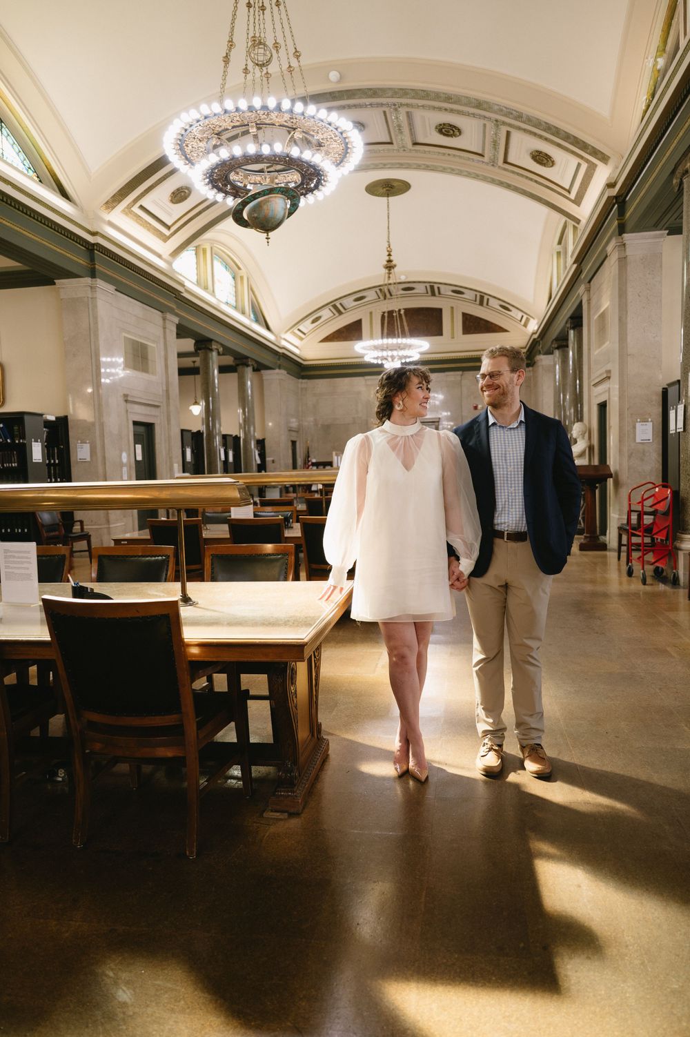 A couple strolls through a grand hall with beautiful chandeliers and classic architecture.