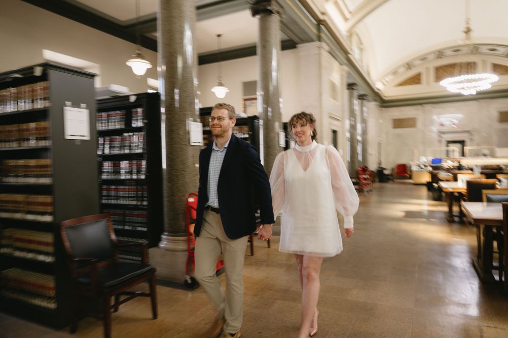 A couple walks through a library space with bookshelves and vintage architecture.