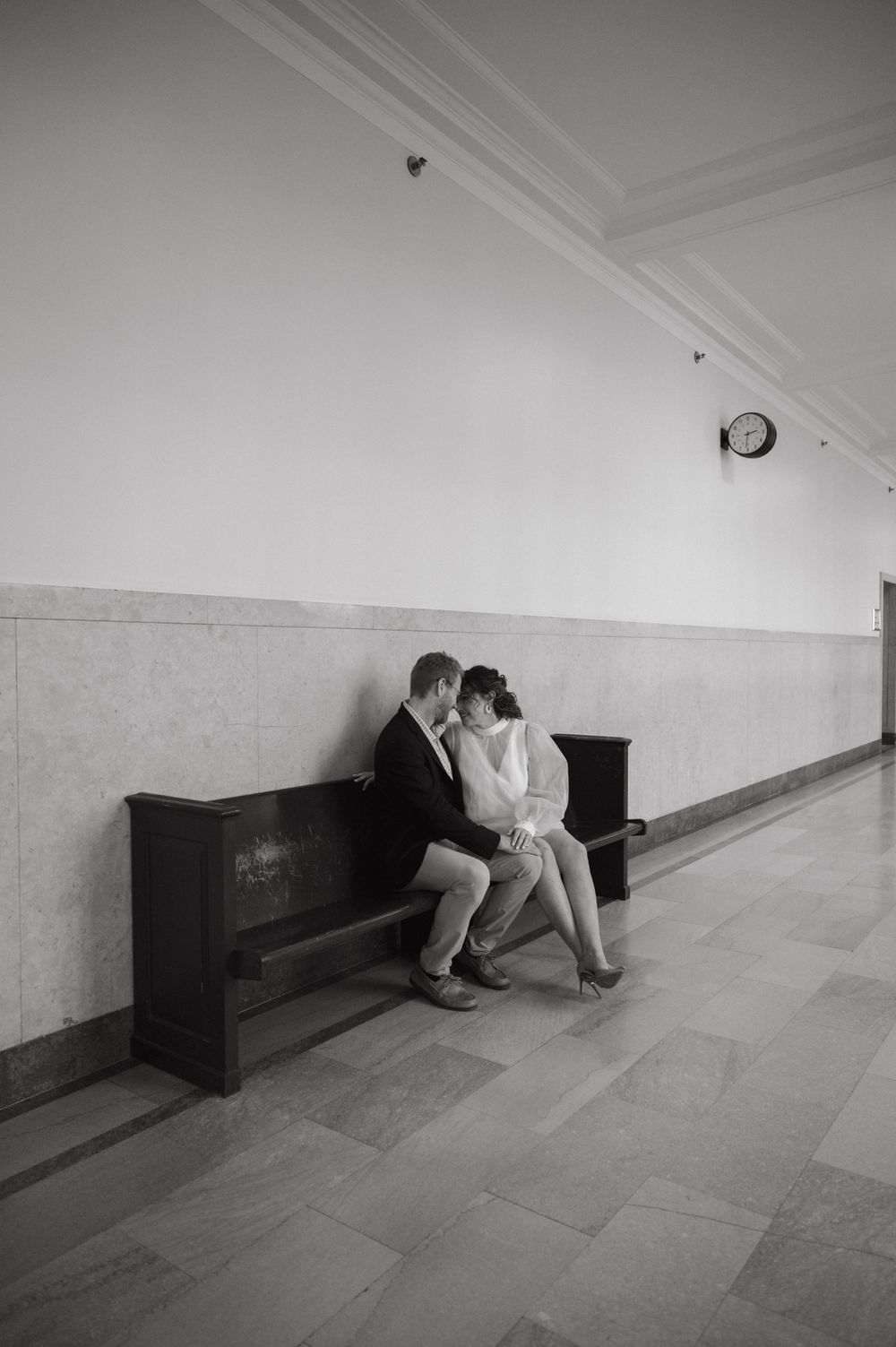 A couple sits close together on a wooden bench in a minimalist hallway.