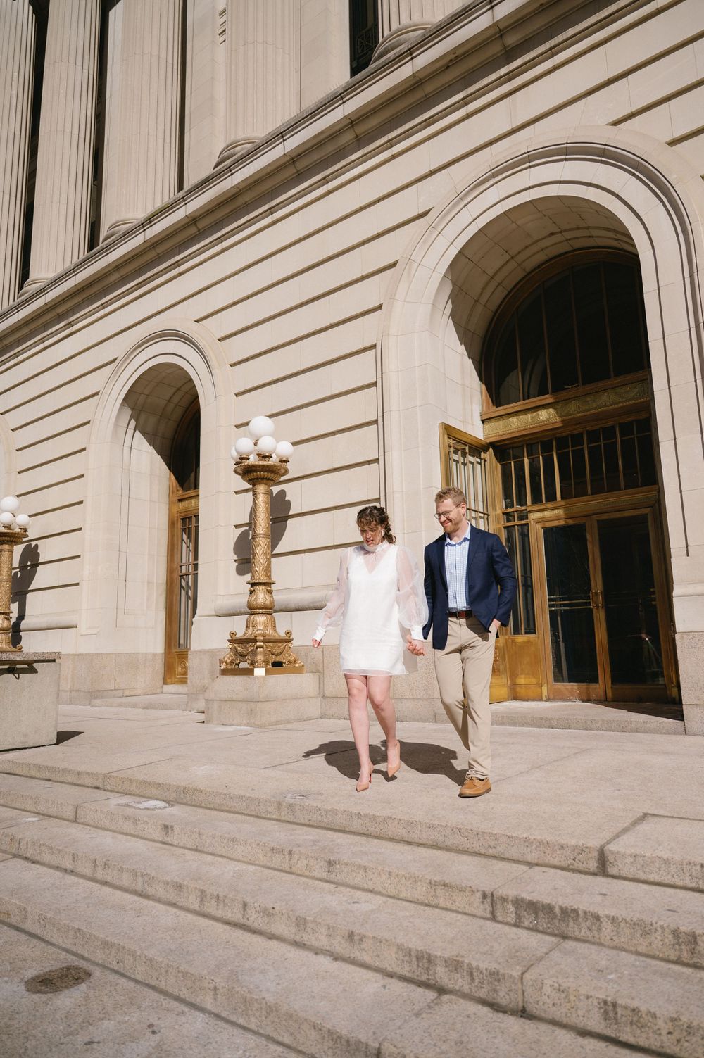 A couple descends grand stone steps of a neoclassical building with arched doorways.