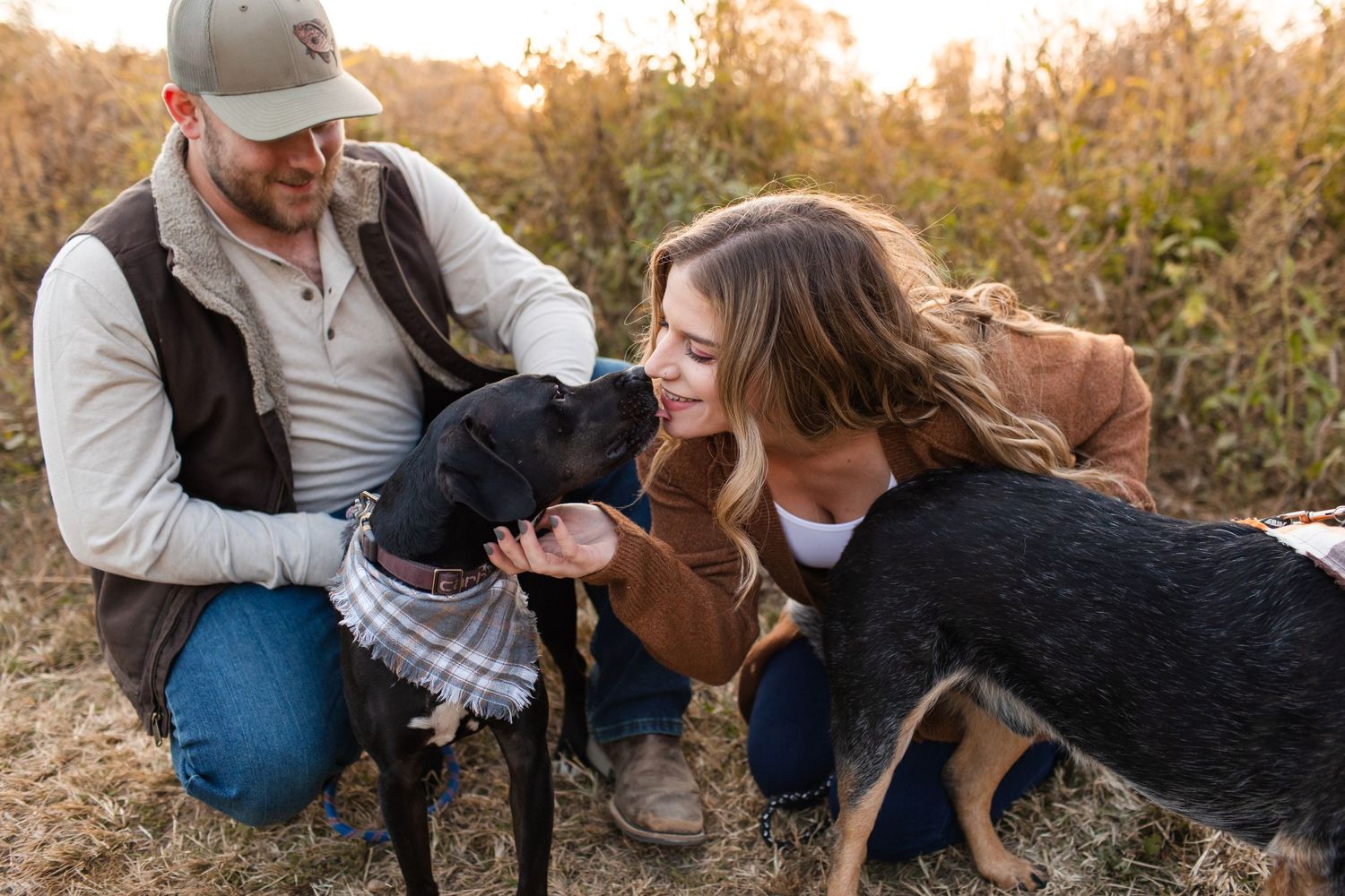 Couple petting their black dogs in a field during sunset.