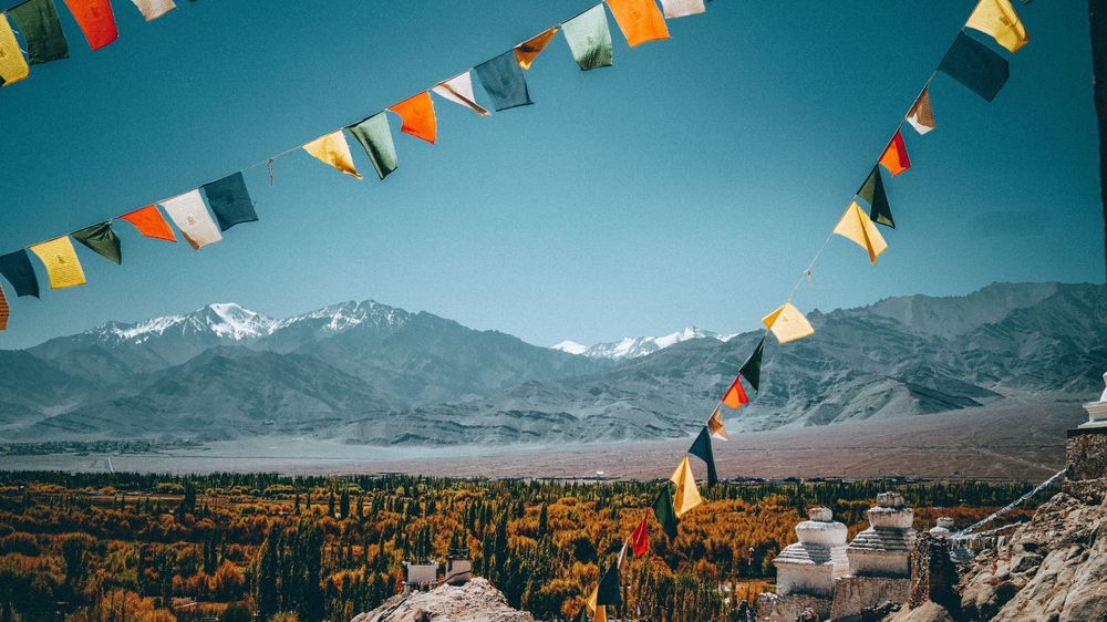 Colorful prayer flags wave against a mountainous landscape with snow-capped peaks in the distance.