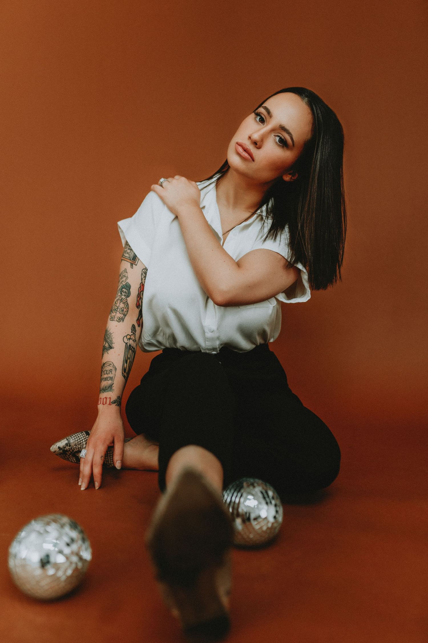 Woman with tattoos sitting on floor in white top and black pants, surrounded by silver disco balls on brown  in Cincinnati
