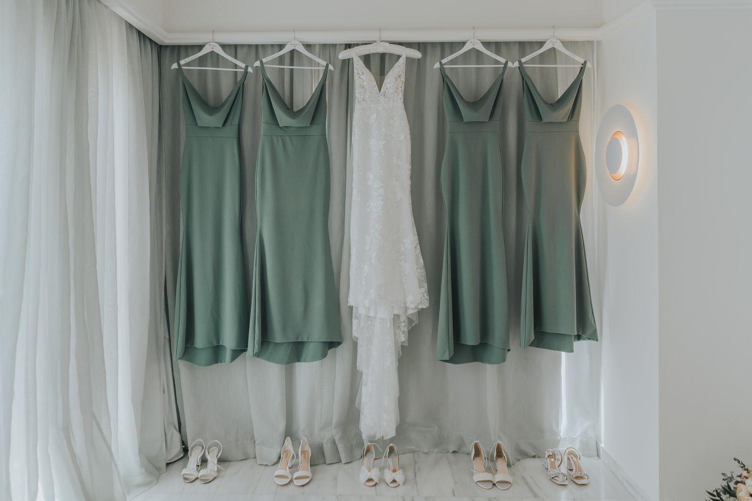Sage green bridesmaid dresses and white wedding gown hanging on display against a white curtained wall.