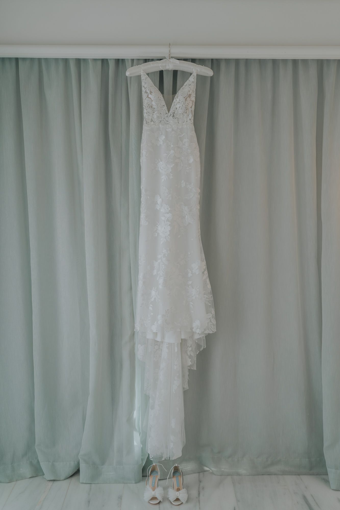 Elegant white lace wedding dress hanging on display against light blue curtains with shoes beneath.